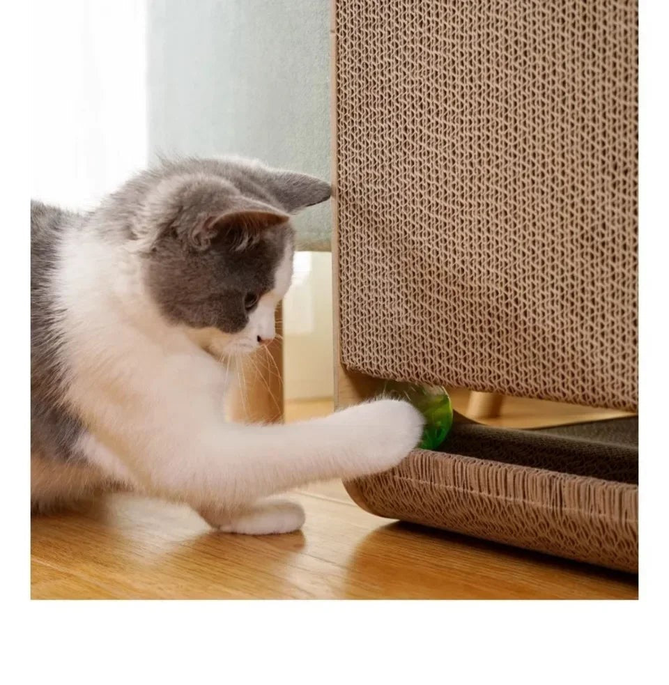 Chat gris et blanc jouant avec une balle verte glissée sous un griffoir vertical en carton ondulé posé sur parquet